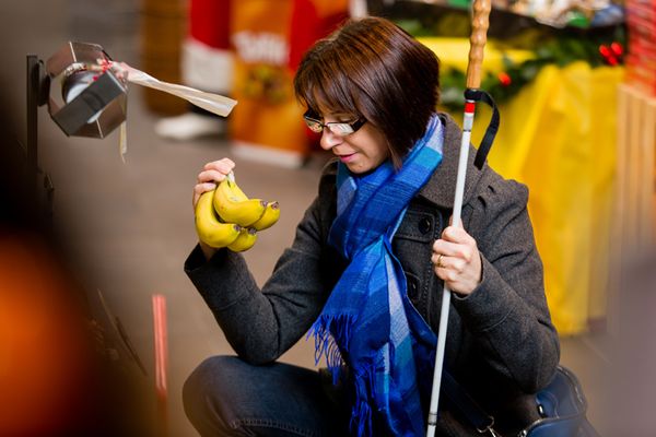 Eine sehbehinderte Frau beim Einkaufen. Sie kniet sich hin, hält sich am weissen Stock fest und versucht, die Preisangaben auf der Etikette an einem Bund Bananen zu erkennen. Bild: SZBLIND