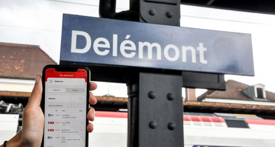 Vor einem Schild der SBB, auf dem die Stadt Delémont angegeben ist, befindet sich ein Smartphone, auf dem die Anwendung geöffnet ist.