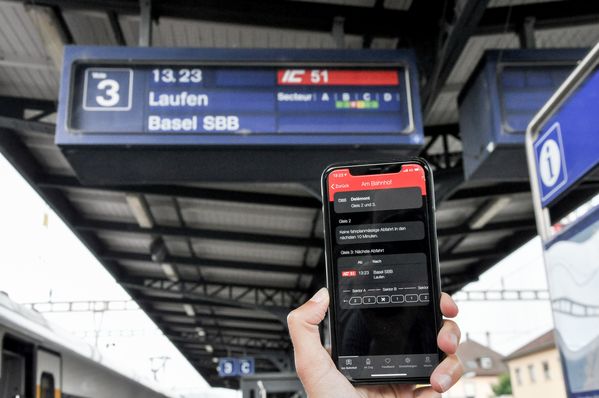 Smartphone vor der Anzeige eines Zuges auf dem Perron, auf dem Bildschirm erkennt man die Zugsinformation