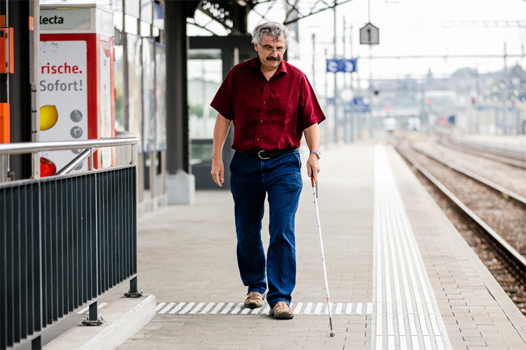 Ein Mann bewegt sich mit seinem Weissen Stock entlang der Leitlinien am Bahnhof. Bild: SZBLIND