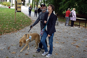 Eine hörsehbehinderte Frau mit Stock und Sonnenbrille geht mit einer freiwilligen Begleitperson und Blindenführhund in einem botanischen Garten spazieren. Bild: SZBLIND