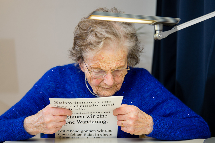 Eine alte Frau liest einen Sehtest an einem Tisch mit einer starken Leuchte. Bild: SZBLIND