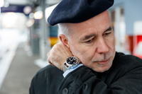 Ein Mann Ende 50 mit einer Barett-Mütze und Anzug steht am Bahnhof und hält sich das Handgelenk mit der ACUSTICA ans Ohr.
