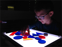 Ein sehbehinderter Junge mit Brille sitzt vor der Lightbox und legt geometrische Figuren auf die beleuchtete Platte.
